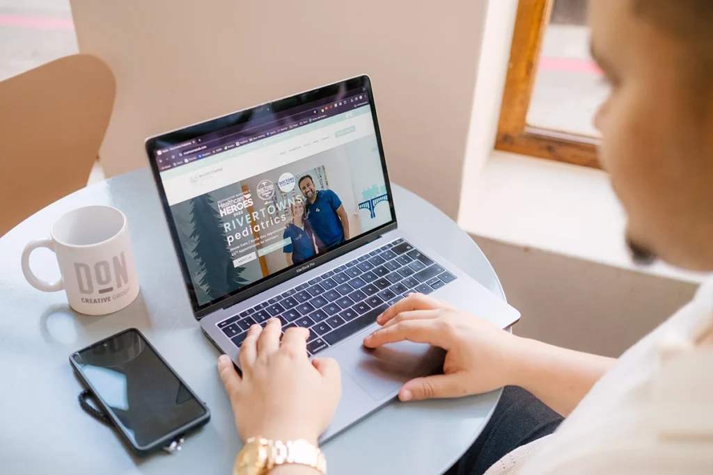 Person browsing the Rivertowns Pediatrics website on a laptop at a round table, with a Don Creative Group mug and phone nearby.