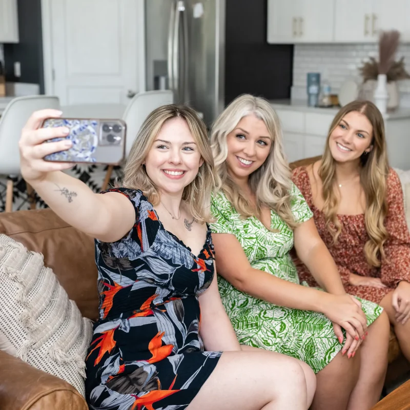 Three smiling women sit on a couch in a modern kitchen taking a selfie together, capturing a fun and casual moment.