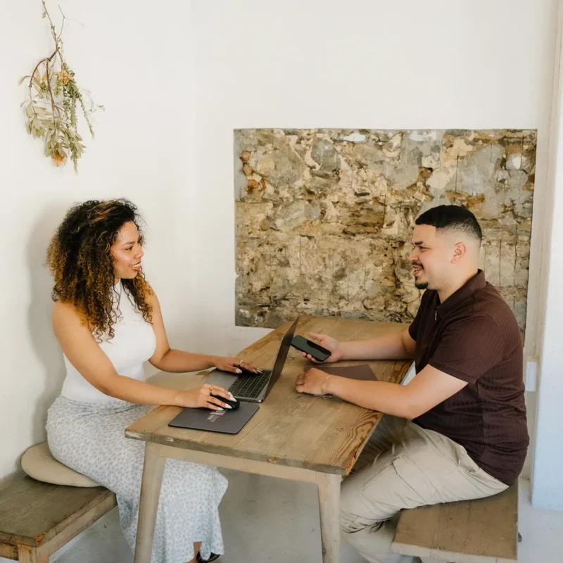 Two professionals collaborating at a wooden table with laptops and a phone in a minimal, modern workspace.