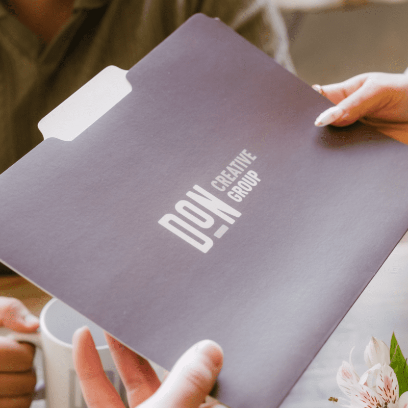 Person handing over a branded Don Creative Group folder during a meeting, with a white coffee mug and flower visible on the table.