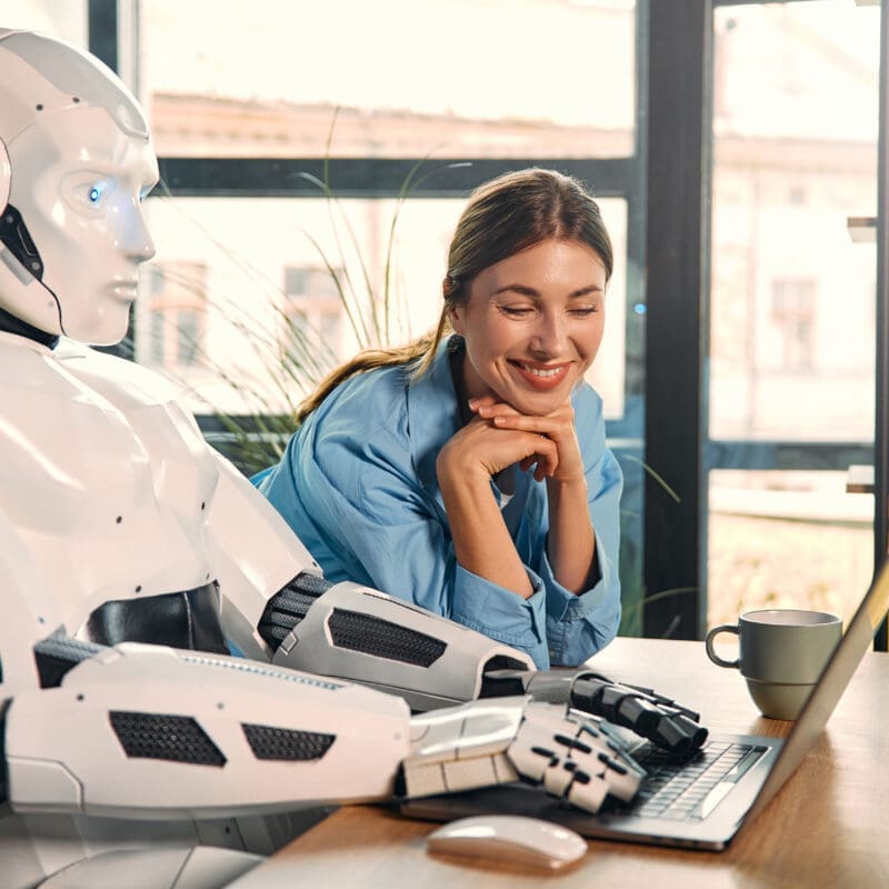 Young woman with a cup of coffee and a humanoid robot working while sitting at a laptop in a modern office. Collaboration between humans and artificial intelligence.