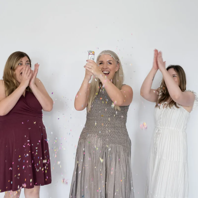 Three women celebrate with colorful confetti, smiling and clapping in front of a white wall, marking a joyful 5th birthday occasion.