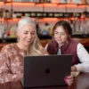 Two women smiling and working together on a laptop in a café setting.