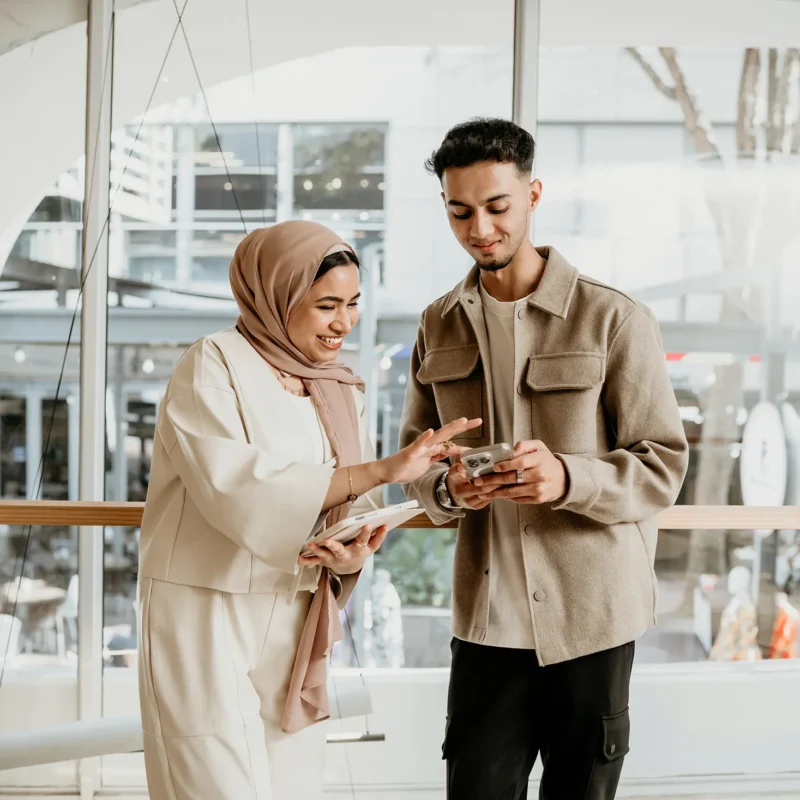Two colleagues smiling and discussing something on a smartphone indoors.