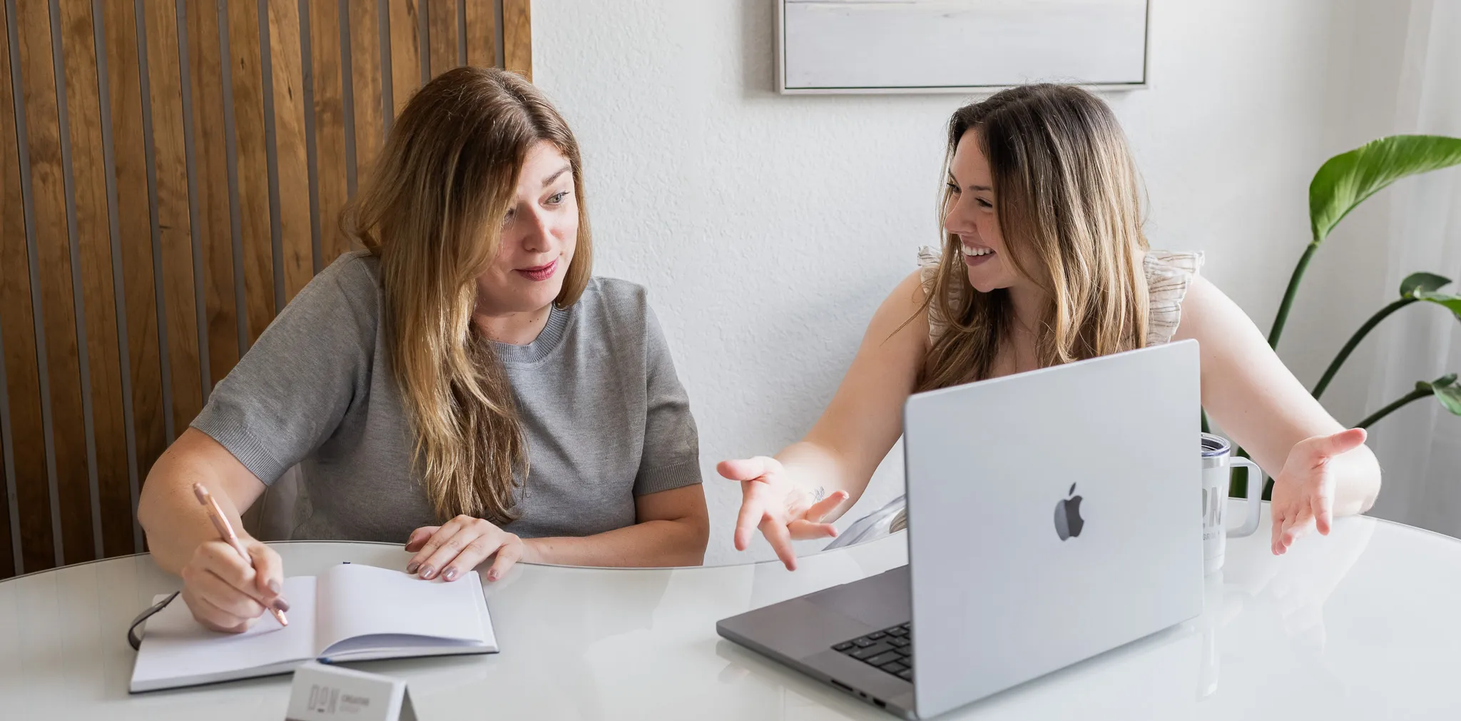 Two creative professionals at Don Creative Group, sit together at a modern white table discussing ideas while working on a laptop and taking notes during a team strategy meeting.