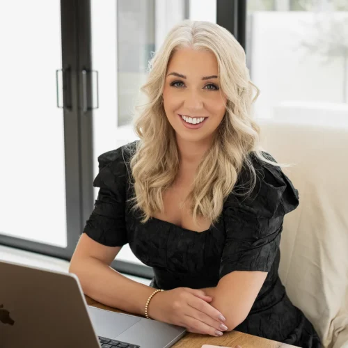 Lauren, a creative professional at Don Creative Group, smiles while sitting at a wooden desk with a laptop and notebook, working on brand strategy in a bright modern office.