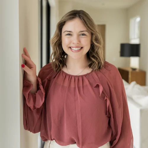 A professional woman with shoulder-length light brown hair, smiles while standing in a bright modern office wearing a rose-colored blouse and white trousers, creating a welcoming and confident team portrait.
