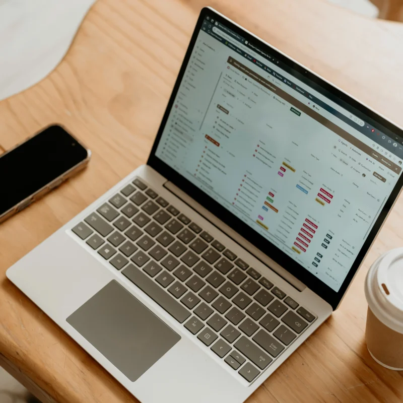 Laptop displaying project management and task tracking tools on a wooden desk beside a smartphone and coffee cup.
