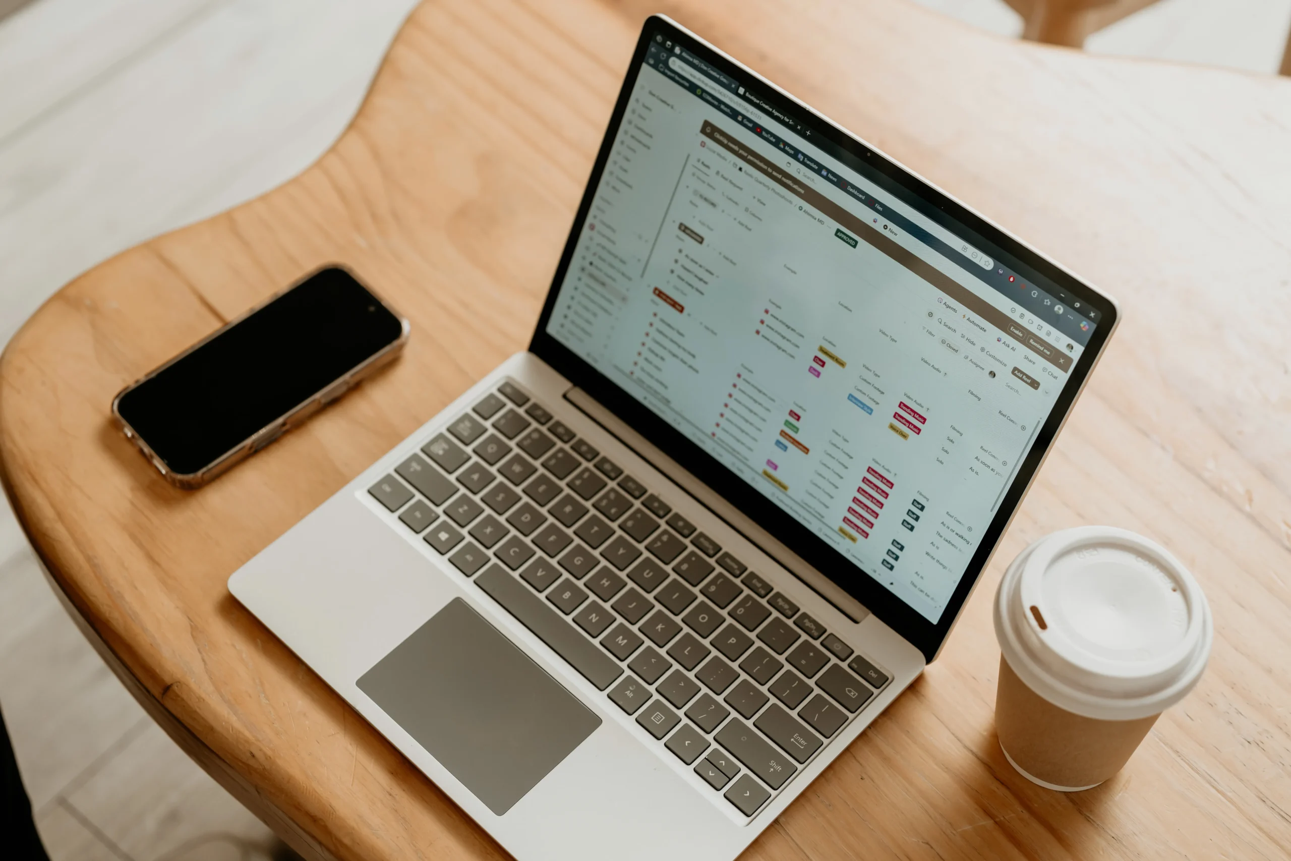 Laptop displaying project management and task tracking tools on a wooden desk beside a smartphone and coffee cup.