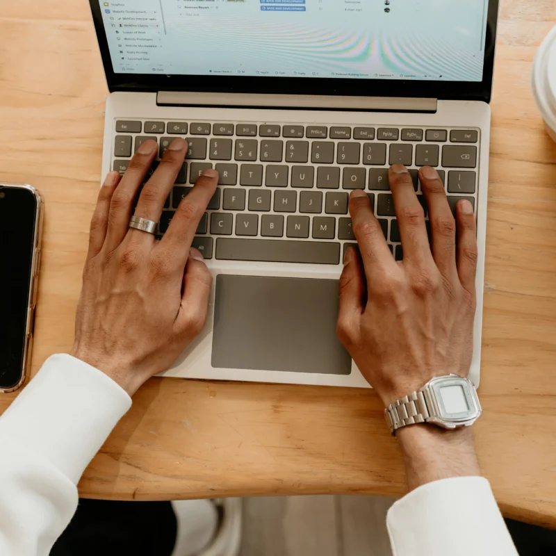 Hands typing on a laptop during website development work, with a phone and coffee nearby on a wooden desk.