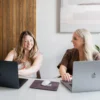 Two women working together at a desk with laptops, smiling during a collaborative meeting at Don Creative Group.