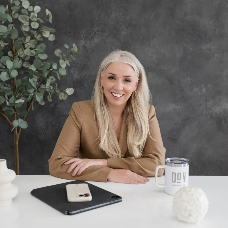 Female creative agency leader smiling at her desk with a laptop, phone, and branded DCG mug, representing Don Creative Group’s professional marketing and design team.