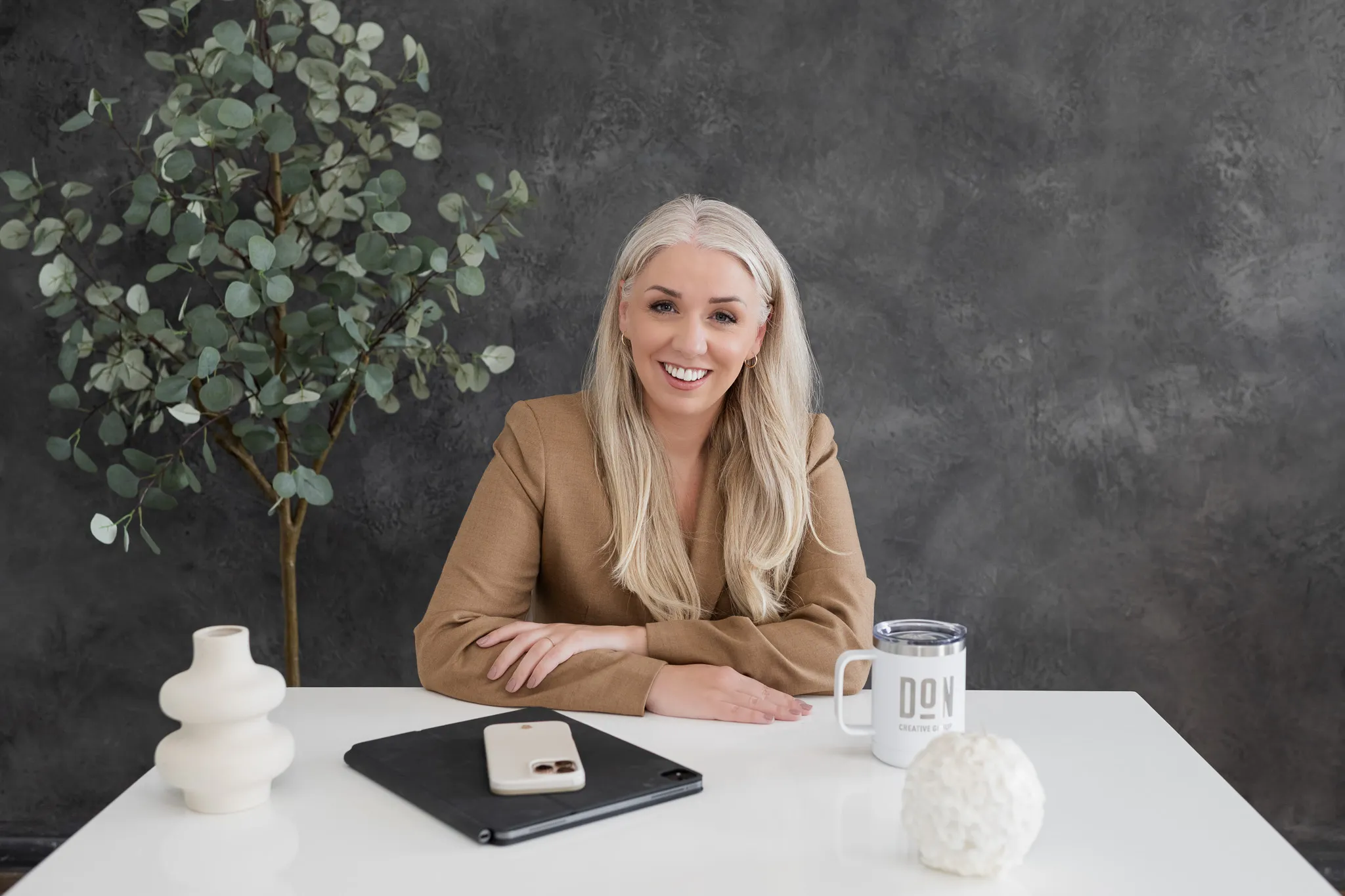 Female creative agency leader smiling at her desk with a laptop, phone, and branded DCG mug, representing Don Creative Group’s professional marketing and design team.