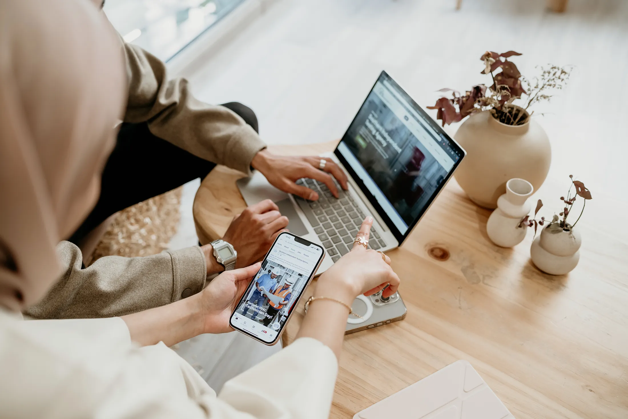 Two professionals reviewing digital content, one using a laptop and the other holding a smartphone, collaborating at a wooden table with minimalist decor.