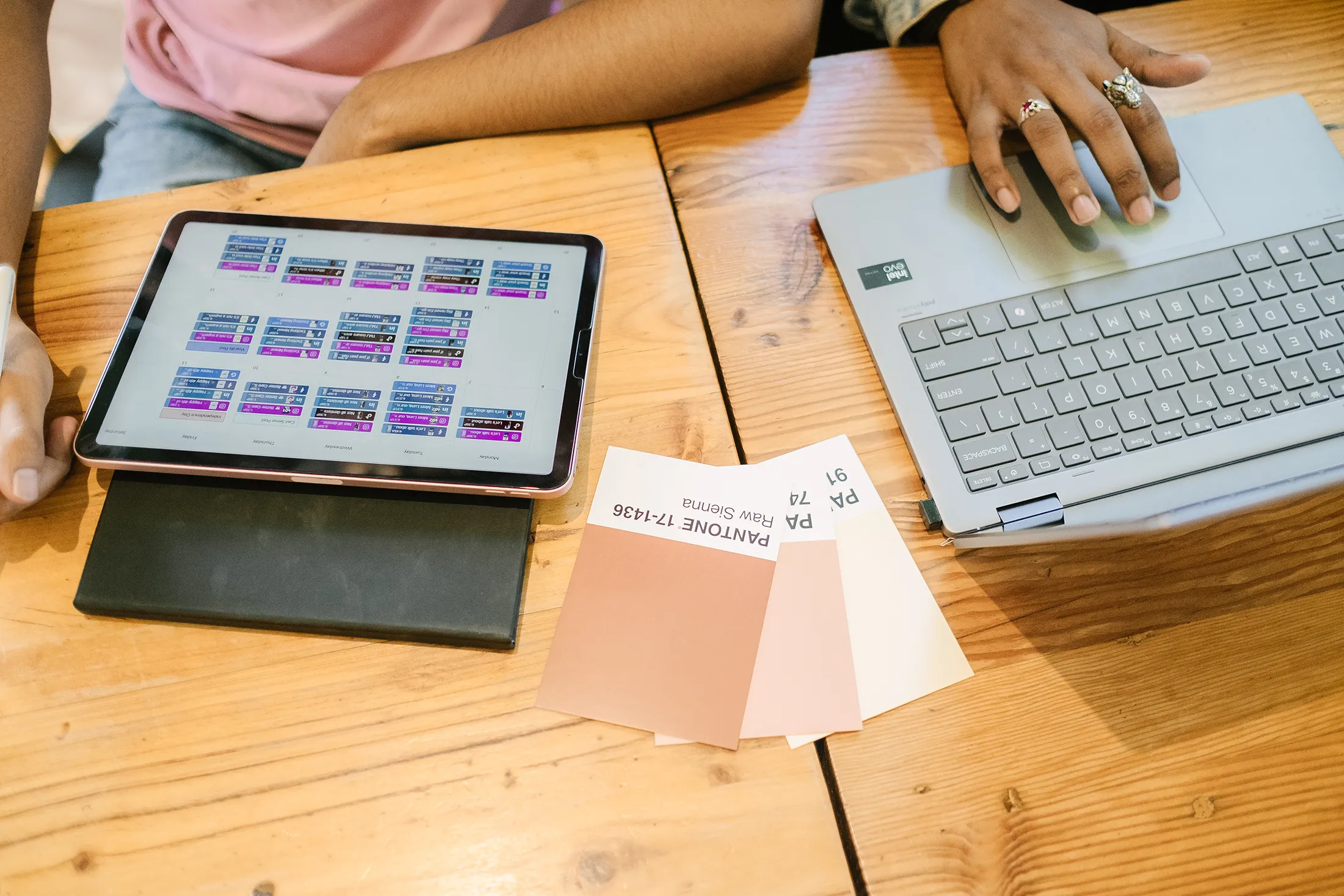 Two people working at a wooden table with a tablet showing a content schedule, a laptop, and color swatch cards laid out for branding and design planning.