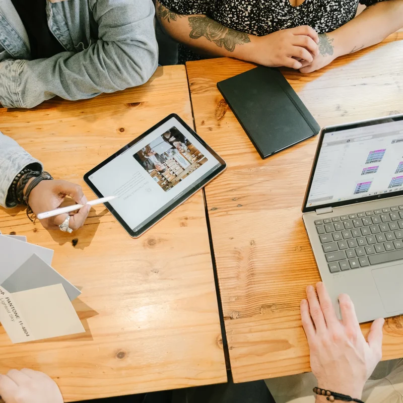Team collaborating at a wooden table with a tablet displaying a website layout, a laptop showing content planning, and Pantone color swatches used for brand identity and rebranding work.