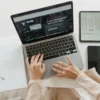 A woman's hands type on a MacBook laptop displaying a social media marketing website, surrounded by a tablet showing the Don Creative Group logo, a smartphone, and an open notebook.