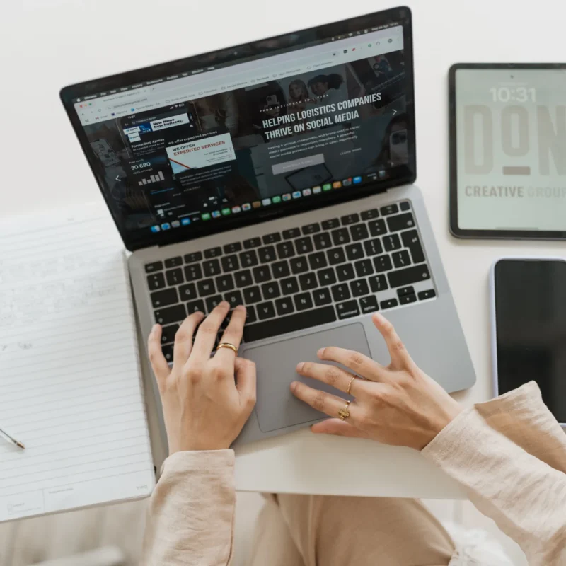 A woman's hands type on a MacBook laptop displaying a social media marketing website, surrounded by a tablet showing the Don Creative Group logo, a smartphone, and an open notebook.