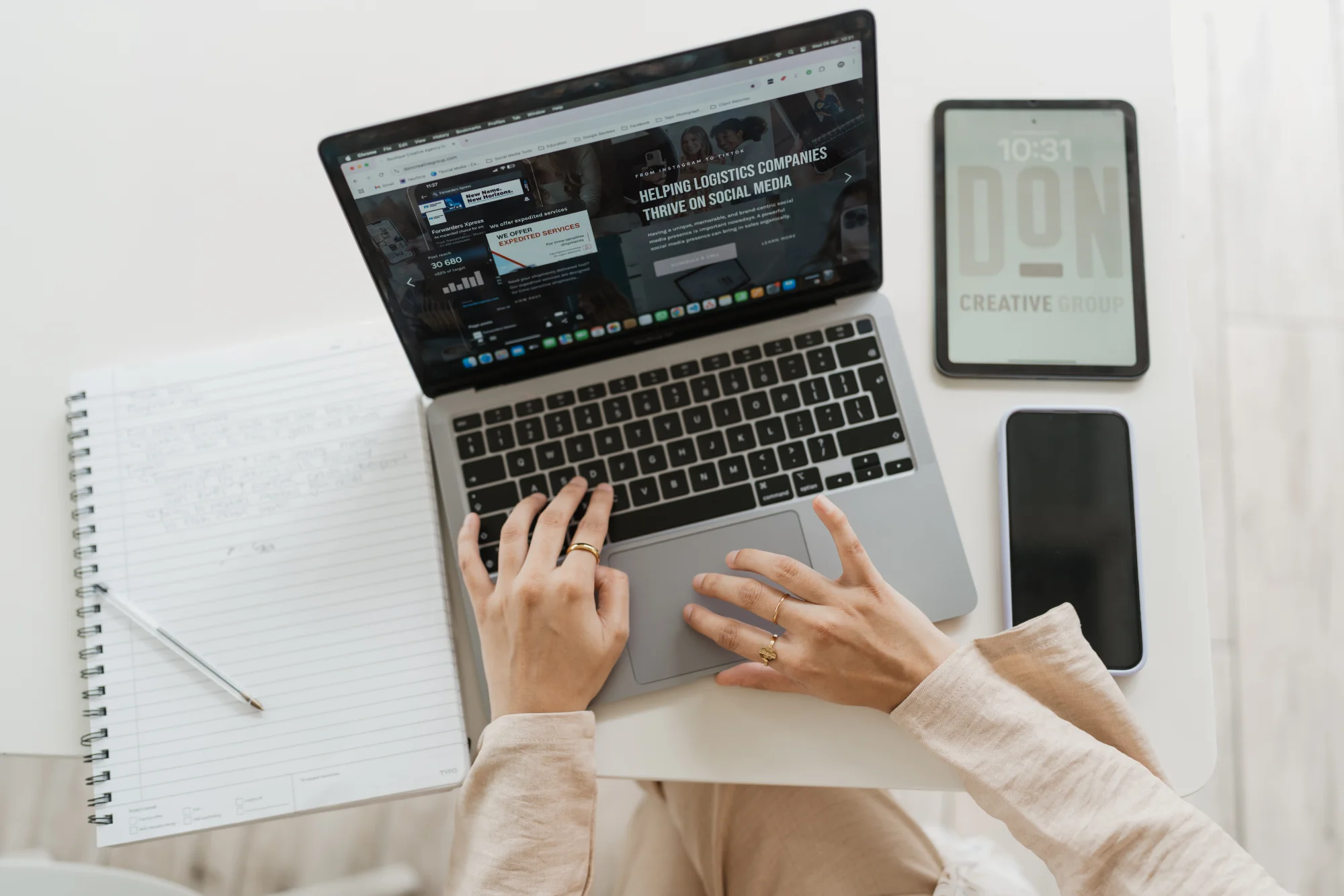 A woman's hands type on a MacBook laptop displaying a social media marketing website, surrounded by a tablet showing the Don Creative Group logo, a smartphone, and an open notebook.