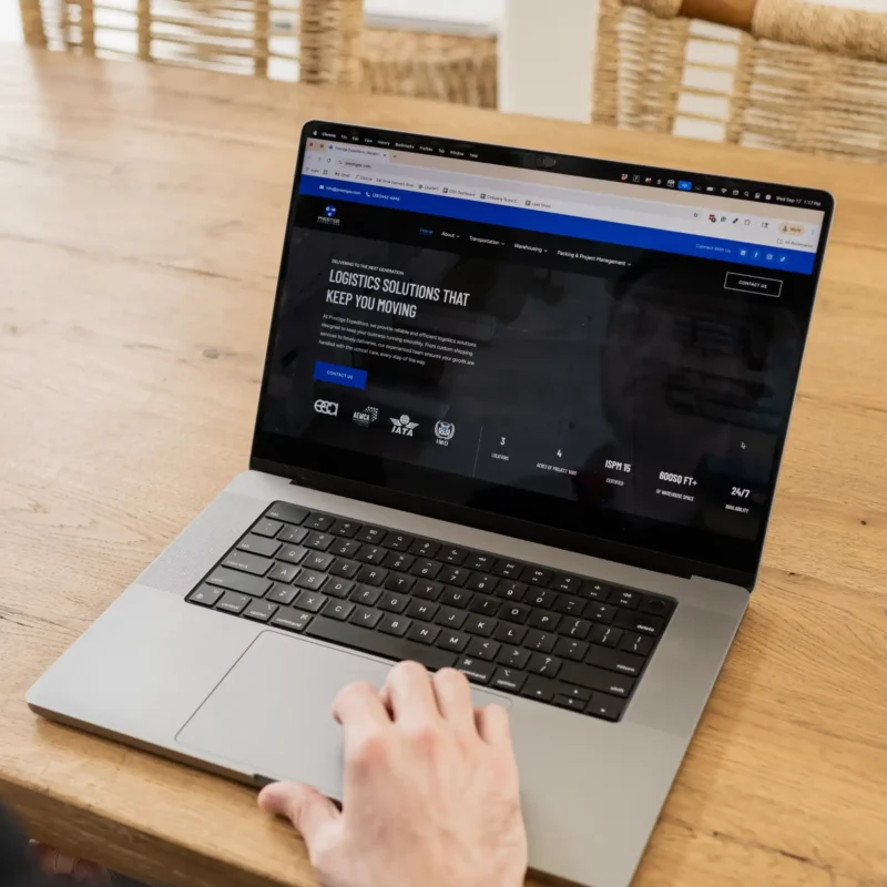 A person browses a logistics company website on a laptop placed on a wooden table in a bright home setting.