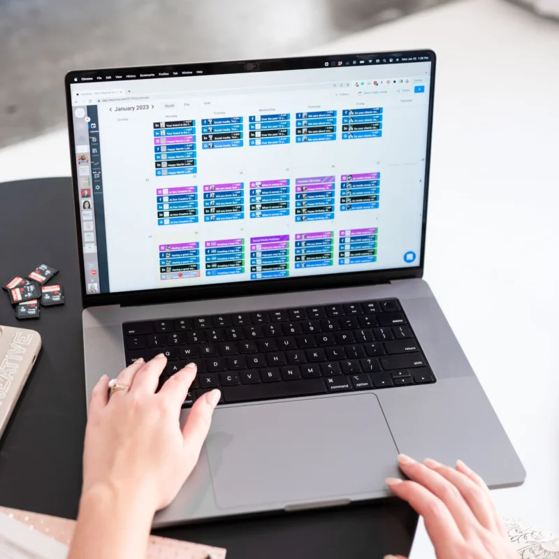 A woman's hands type on a MacBook laptop displaying a colorful social media content calendar for January 2023, with SD cards and a notebook visible nearby.