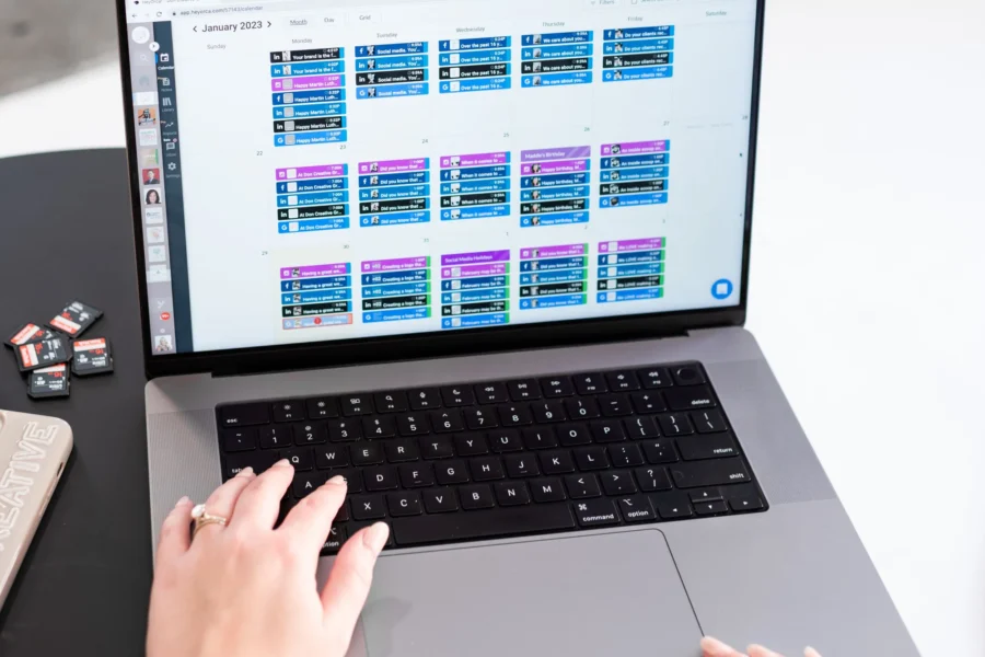 A woman's hands type on a MacBook laptop displaying a colorful social media content calendar for January 2023, with SD cards and a notebook visible nearby.