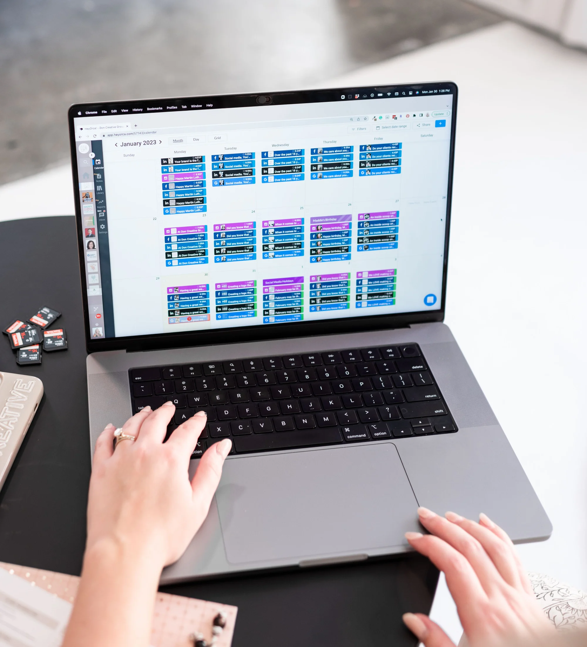 A woman's hands type on a MacBook laptop displaying a colorful social media content calendar for January 2023, with SD cards and a notebook visible nearby.
